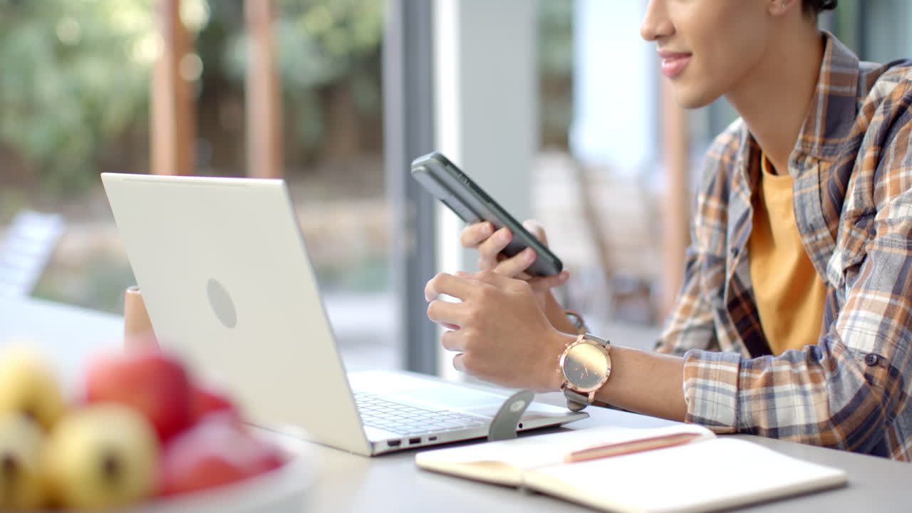 Using smartphone and laptop, young teenage boy working from home office desk