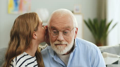 Close-Up-Of-The-Young-Blond-Teen-Girl-Whispering-To-The-Grandfather's-Ear-In-The-Cozy-Living-Room