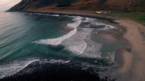 Vista-Aérea,-órbita,-De-Las-Olas-Rompiendo-En-La-Playa-De-Unstad,-Mientras-Los-Surfistas-Disfrutan-De-La-Hermosa-Puesta-De-Sol,-En-Un-Día-De-Cielo-Azul-Claro,-Islas-Lofoten,-Noruega