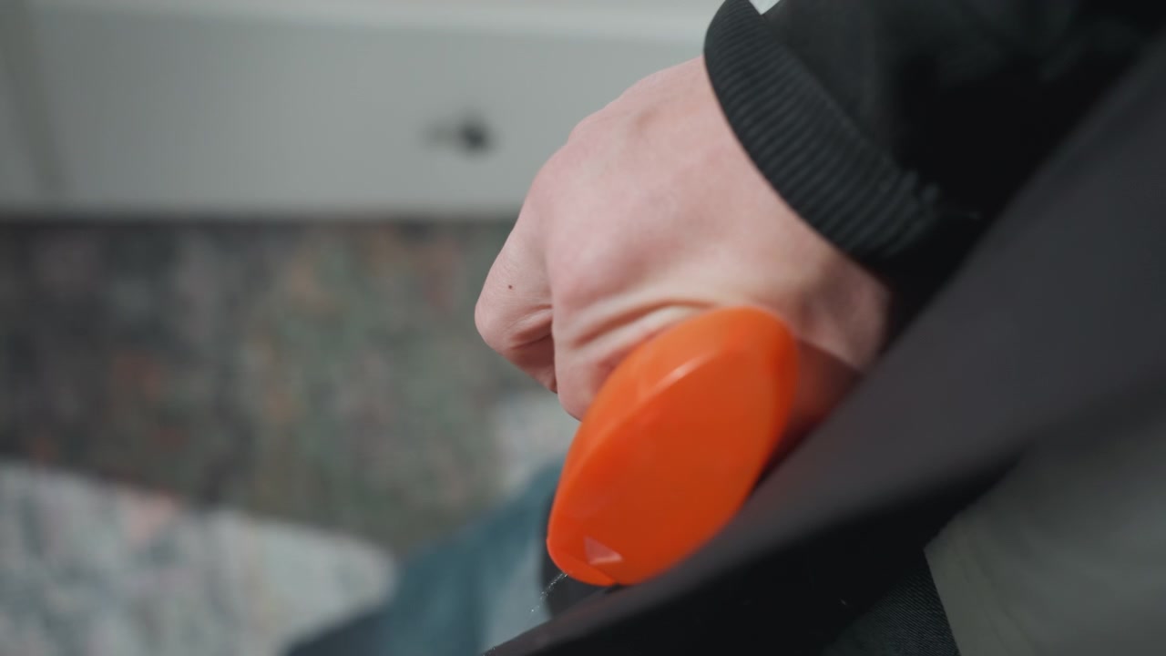 Mano usando rodillo quitapelusas naranja en el bolsillo de un abrigo, con fondo de alfombra interior. Primer plano de eliminación de bolitas de tela, propietario ordenando el armario, movimiento suave y detalle texturizado para limpieza del hogar y cuidado de prendas.