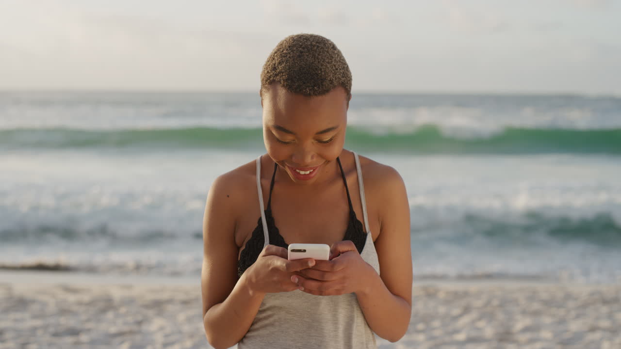 giovane donna afroamericana che usa lo smartphone sulla spiaggia, che scrive messaggi, che naviga online, che si gode la comunicazione mobile in un mare soleggiato