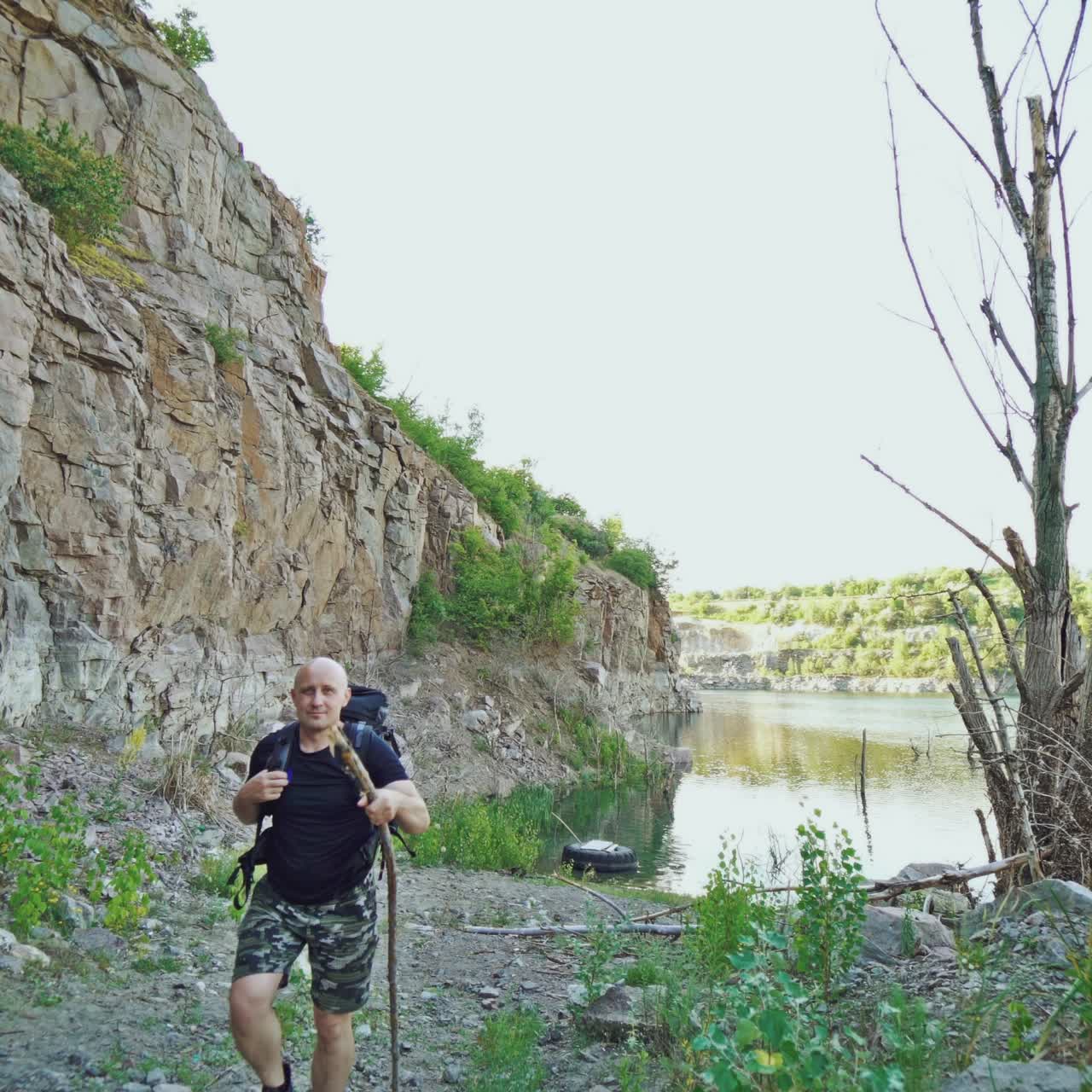 adult tourist is walking along the bank of the lake with a stick in his hand and with a backpack on his shoulders on the background of high rocks in the summer