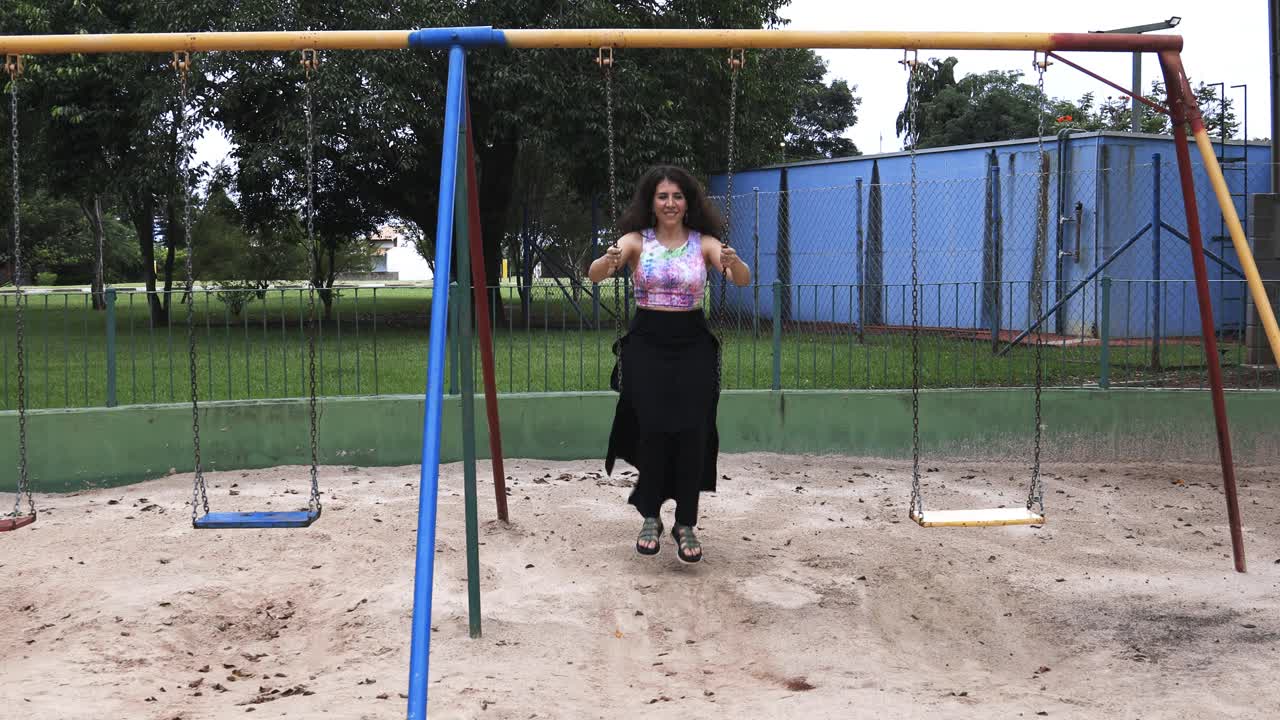 mujer joven con falda larga y camisa colorida balanceándose y riéndose sintiéndose libre y feliz en un parque visto desde el frente con un sutil movimiento de cámara