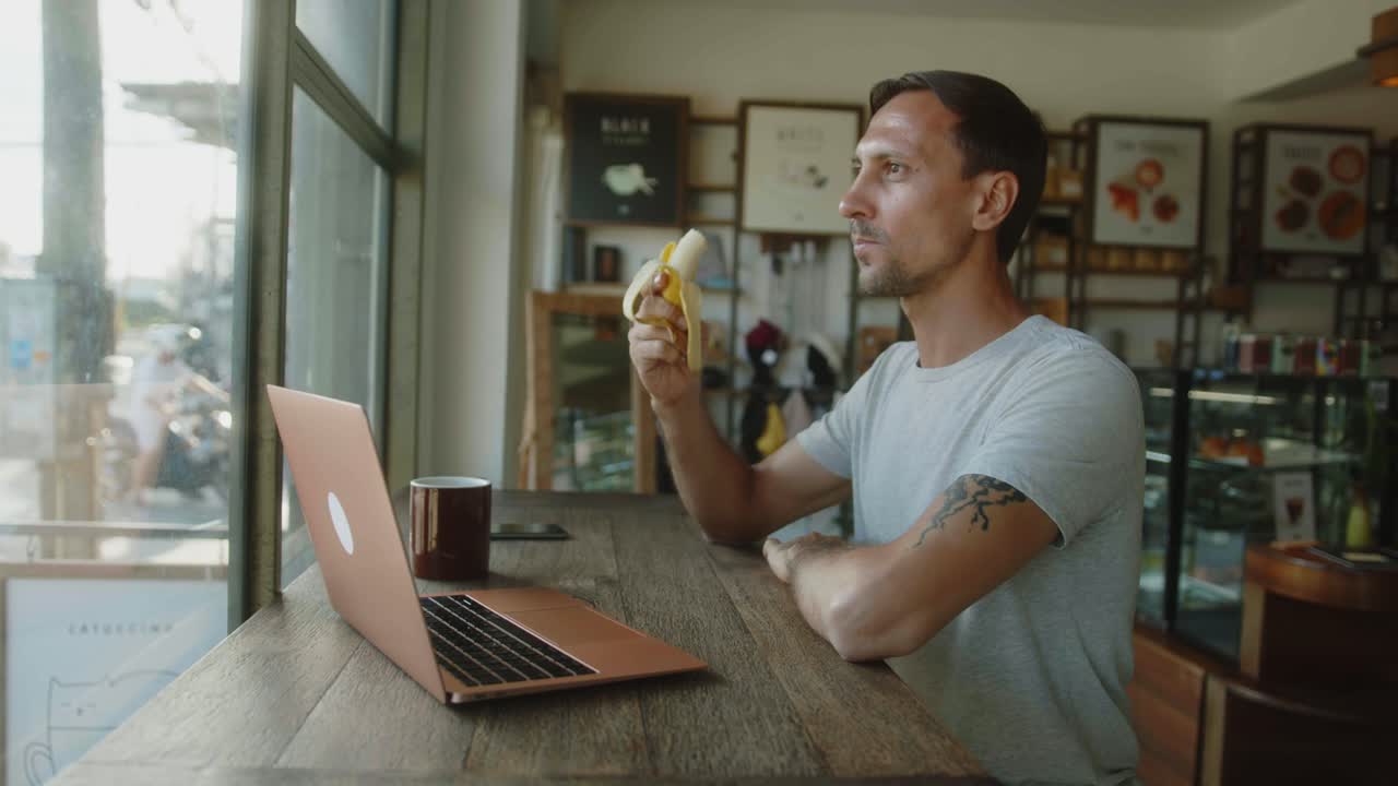 Man eating banana in a coffee shop while working on a laptop