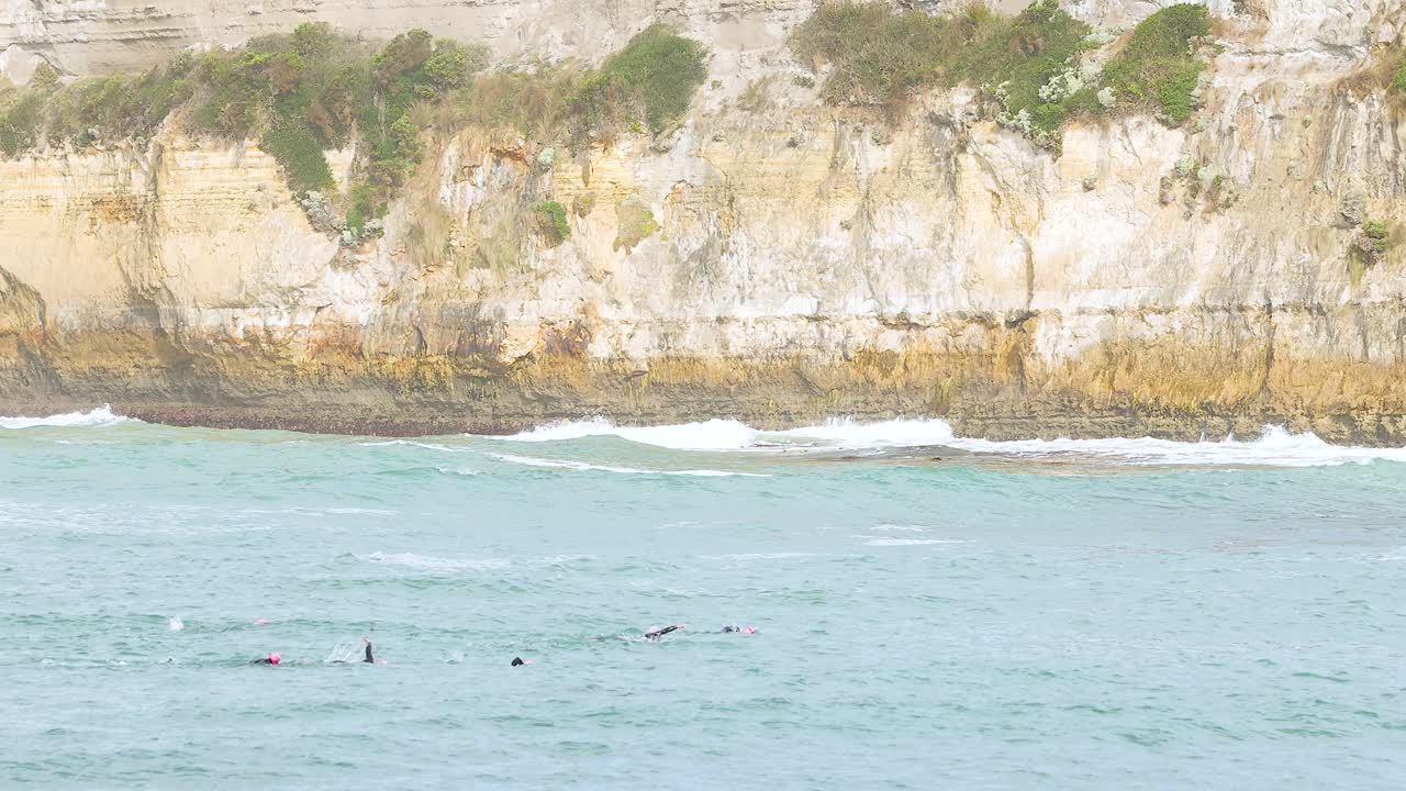 Swimmers traverse turquoise waters near rocky cliffs under bright daylight at Port Campbell, Australia. The scene captures movement and natural beauty