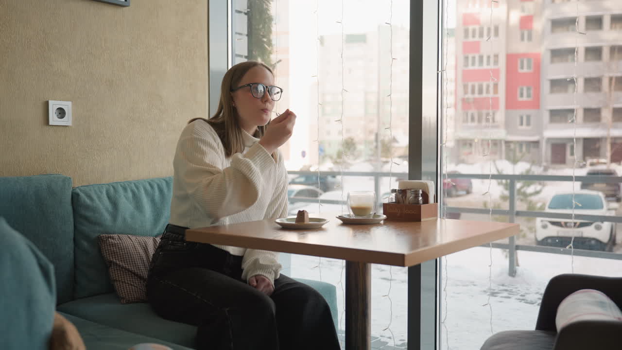 young lady in white sweater enjoys breakfast alone in bright modern cafe, slicing chocolate dessert with fork while seated near large window overlooking snowy street scene and parked vehicles