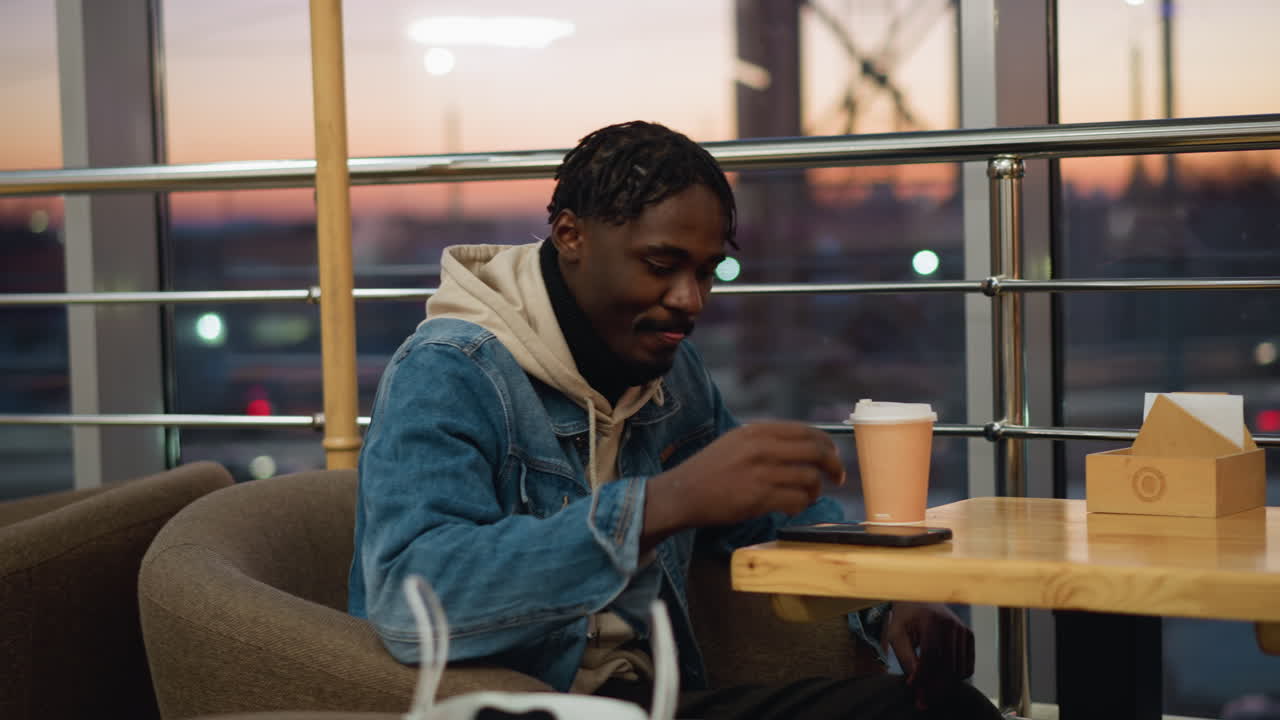 Young man in denim jacket and beige hoodie sits comfortably in modern cafe during sunset, gently sipping coffee with relaxed posture, surrounded by warm tones, armchairs, and soft glowing twilight behind windows