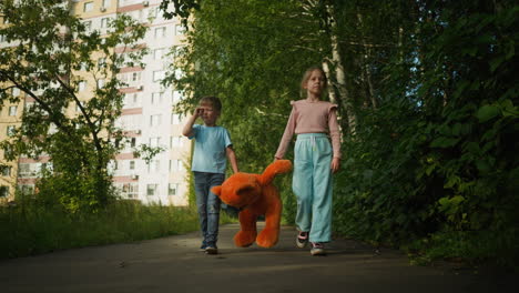 children walking together on paved path bordered by greenery and apartments, with boy inspecting object using small microscope and girl lifting large teddy bear to keep it from touching ground