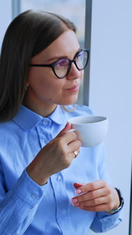 Young lady with long hair drinking coffee standing and looking into window. Relaxed calm female enjoying view from window. Vertical video