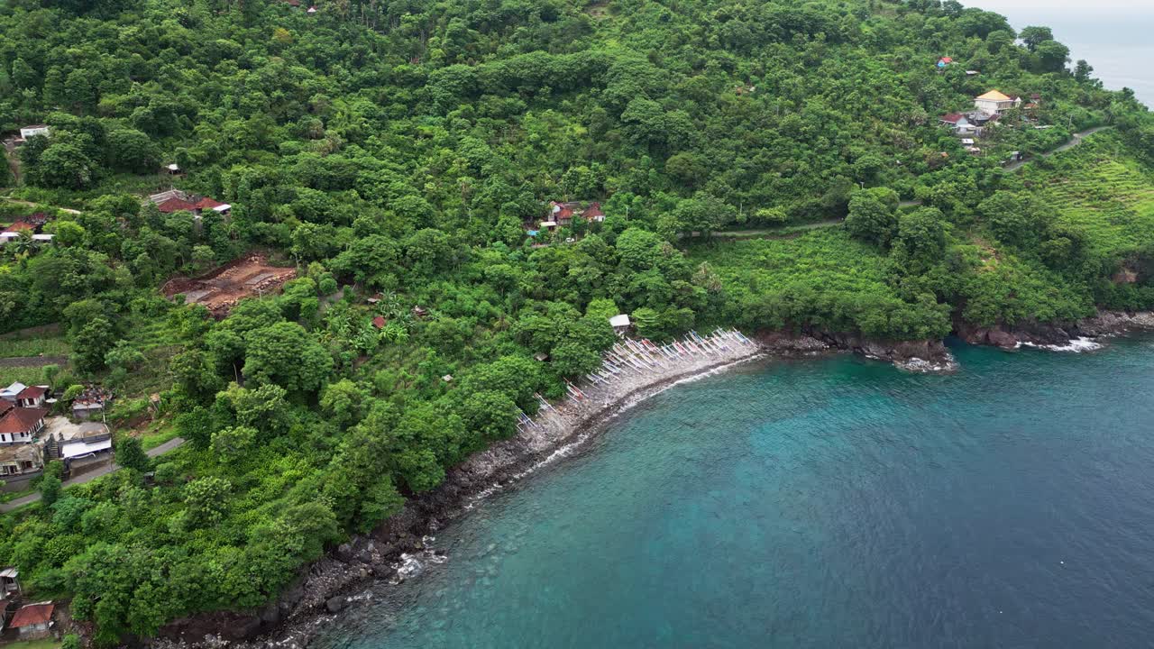 Riprese aeree con drone mostrano una piccola città costiera a Bali delimitata da fitte colline tropicali con file di barche da pesca locali allineate lungo una costa curva e acqua cristallina dell'oceano.