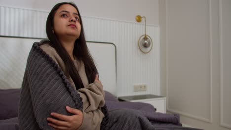 mujer triste con un resfriado en una chaqueta marrón y una estufa gris oscuro toma pastillas durante su enfermedad sentada en la cama en un apartamento moderno en el dormitorio
