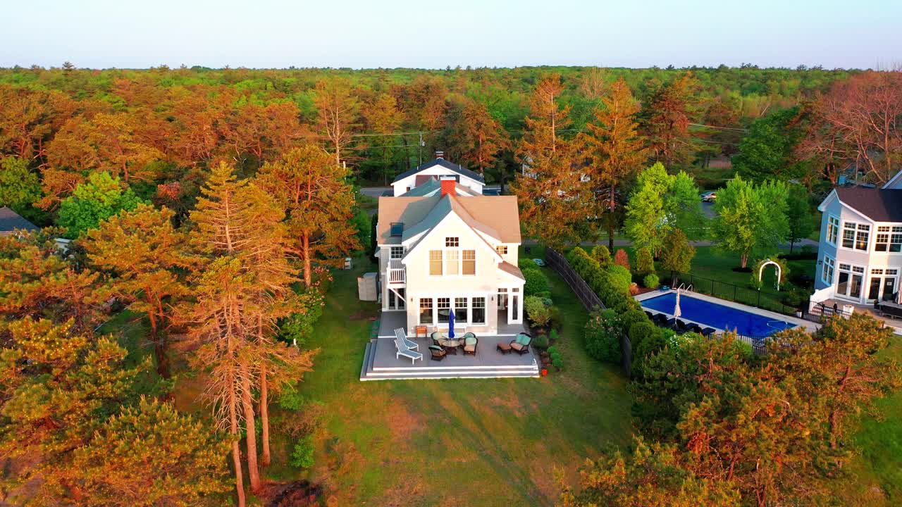 Aerial drone real estate video of a charming suburban cottage home featuring a spacious back porch, inviting patio furniture, large yard, and surrounded by vibrant green trees and lush landscape