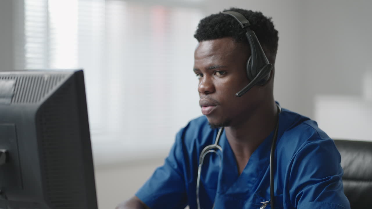 Afroamerikansk mannlig lege eller sykepleier med hodesett og datamaskin jobber på sykehus. Ung profesjonell terapeut/lege konsulterer kunde/klient ved hjelp av fjernkommunikasjon og snakker via webkamera.