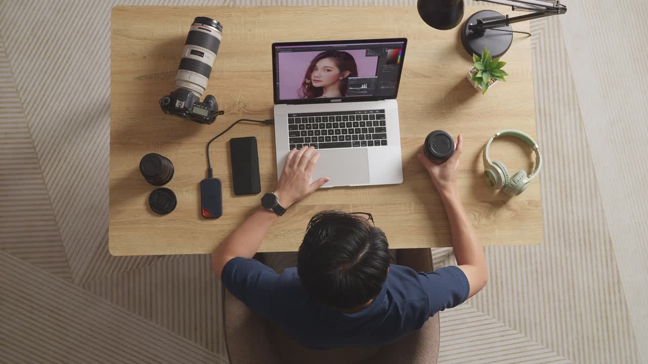 Top View Of Asian Male Editor Drinking Coffee While Sitting In The Workspace Using A Laptop Next To The Camera Editing Photo Of A Female At Home