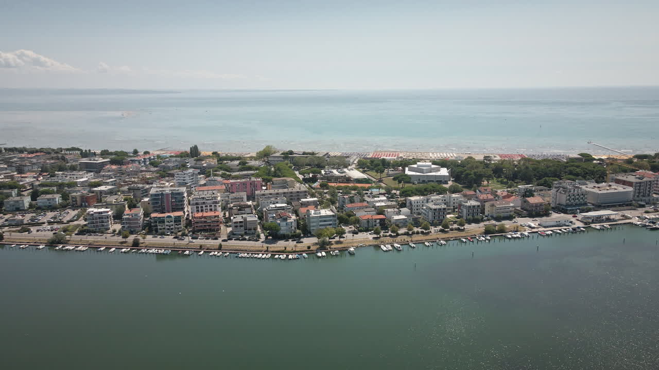 Panoramica fluida di un drone 4K UHD verso sinistra sul lungomare di Grado, che cattura edifici residenziali, barche ormeggiate nella laguna e il Mare Adriatico all'orizzonte – ideale per film di viaggio