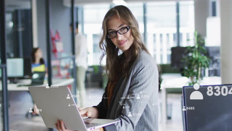 professional woman holding laptop while business user-icon graphics floating across office monitors