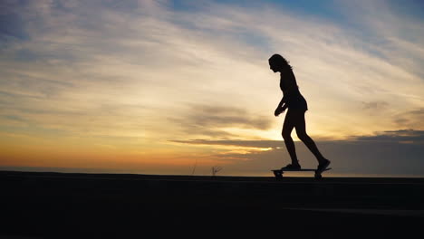 Against-the-stunning-mountainous-backdrop-and-a-captivating-sky,-a-woman-enjoys-slow-motion-skateboarding-on-a-road-at-sunset.-She's-wearing-shorts.