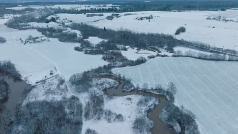 vista aérea del valle del río abava en un día nublado de invierno, campos cubiertos de nieve, el río abava lleno de aguas de inundación oscuras, amplia toma de drone moviéndose hacia adelante, inclinándose hacia abajo