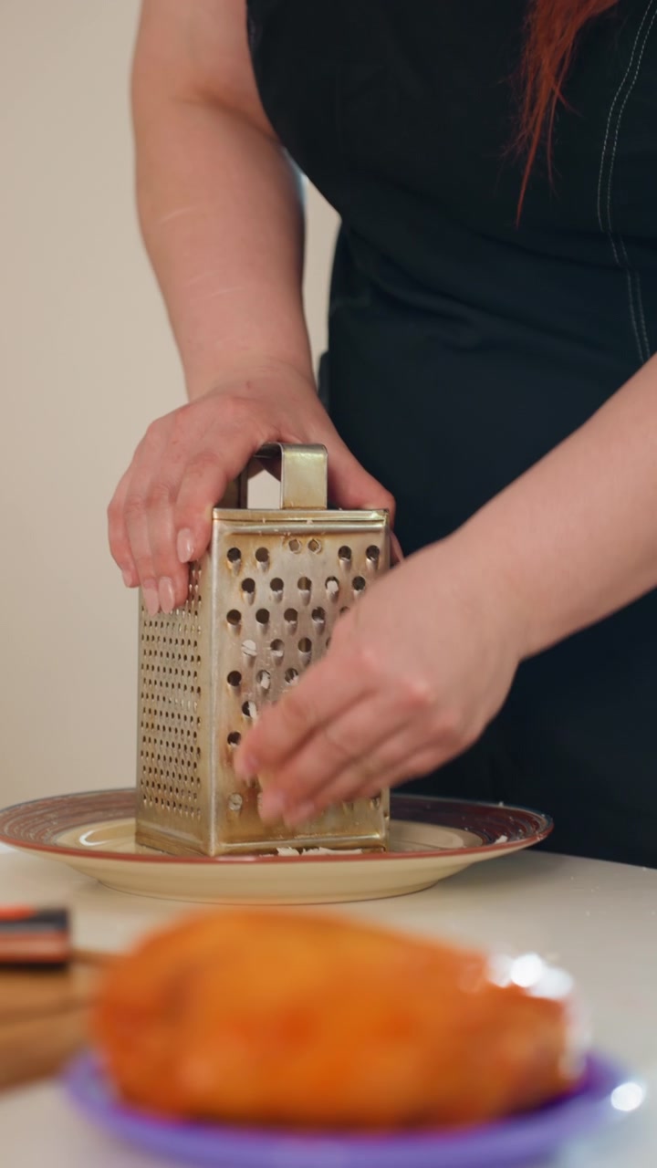 Close view of hands using metal box grater to shred food over plate while shavings collect below; person in dark top works at kitchen table beside blurred baked pastry, preparing fresh topping