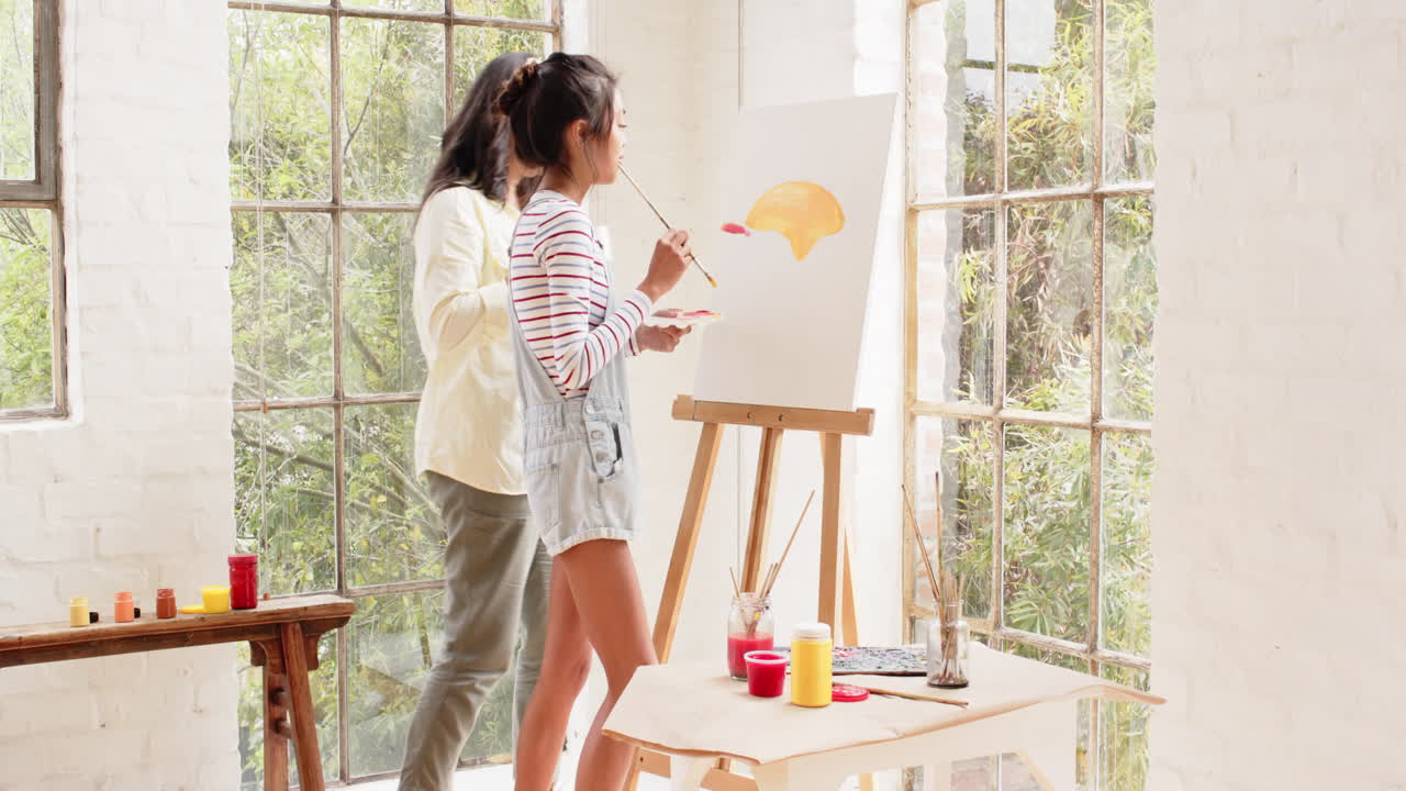 Multiracial young woman painting with grandmother at home, sharing creative moment together