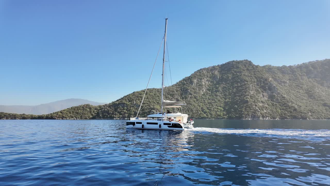 Turkey, Marmaris, a catamaran is entering the Marmaris Bay on a sunny day with no clouds, splashes behind the boat, two people on the top deck.