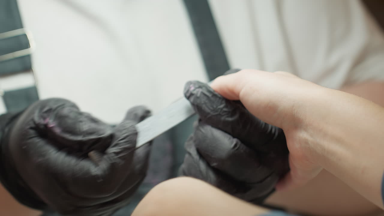 Nail technician wearing black gloves precisely filing customer's fingernail using hand filer during manicure session, both hands clearly visible in close up with customer receiving nail care