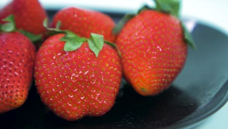 Fresh-big-red-tasty-ripe-strawberries-rotates-slowly-on-a-black-plate-on-light-blue-background,-healthy-food-concept,-extreme-close-up-shot,-camera-rotate-right