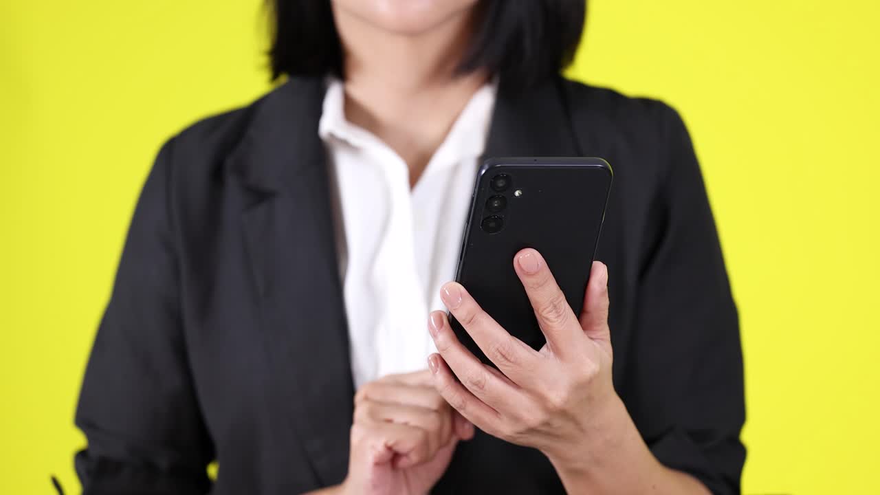 Donna in blazer nero interagisce con uno smartphone, in piedi, su uno sfondo giallo brillante, illuminazione da studio uniforme.