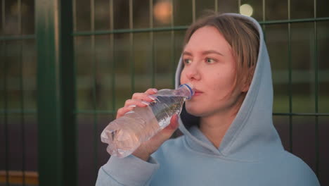 young lady in cyan hoodie hydrates by sipping water from a bottle, seated outdoors with a blurred bar fence background, capturing a peaceful recovery moment after exercise