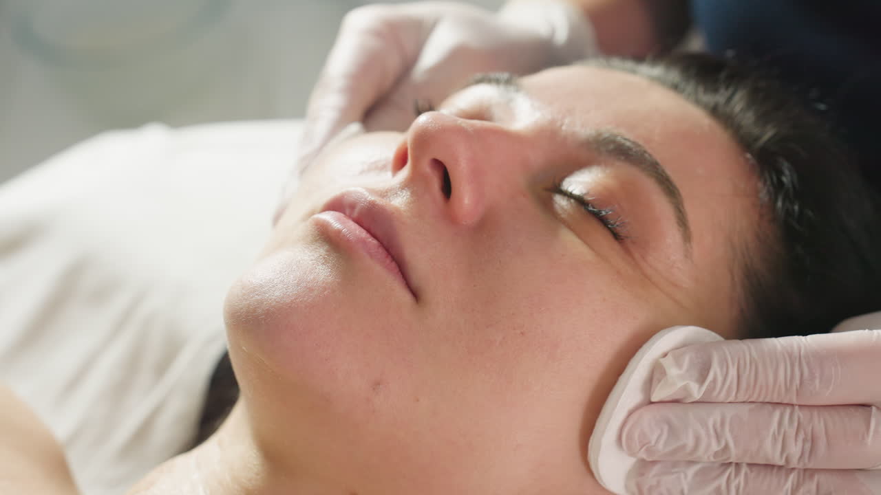Close up of therapist hands wiping patient face with cotton pads for facial detoxification during gentle spa treatment session under soft ambient lighting in bright clinic environment