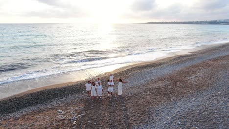 zoom out of group of multigenerational asian family waving smiling standing at sunset on picturesque mediterranean sea shore. gorgeous landscape at subtropical tourist resort with happy people resting.