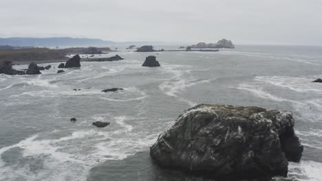 Stormy-day-aerial-of-a-rocky-ocean-shore,-tracking-over-a-large-rock-in-the-ocean,-waves-crash-against-dark-sea-rocks,-the-sky-looks-dark-with-clouds