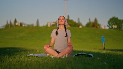 a young woman settles onto her yoga mat, taking a deep breath as the golden evening sun gives her skin a beautiful glow. a green hill rises behind her as the camera moves gently to the left.