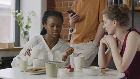 two multiracial female roommates having breakfast and talking together in the kitchen at home