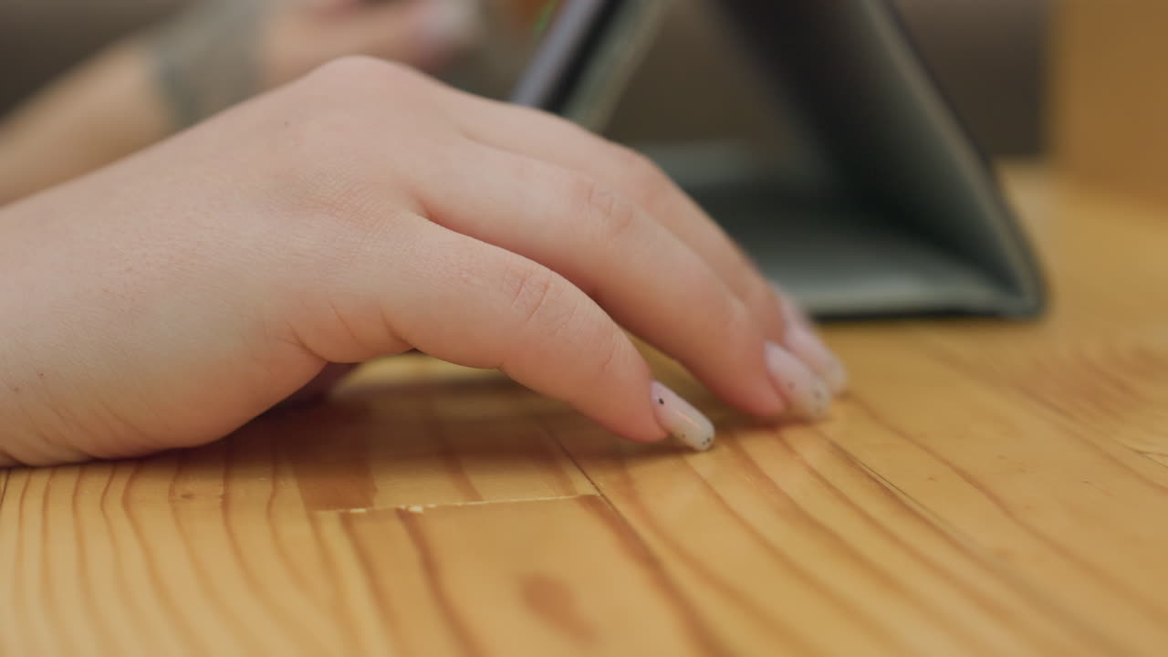 close up of fair skinned female hand with manicured nails gently tapping on wooden table surface near tablet in calm indoor setting with relaxed mood and soft light reflecting natural tones