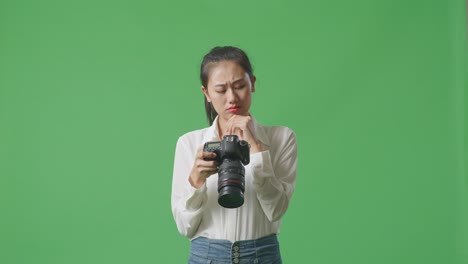 asian photographer thinking about the pictures footage and raising her index finger while looking at the pictures in the camera on green screen background in the studio