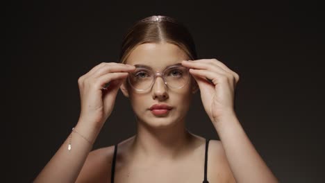 Plano-Medio-De-Una-Mujer-Joven-Y-Bonita-Que-Necesita-Ayuda-Visual-Porque-Su-Vista-No-Es-Lo-Suficientemente-Buena-Y-Se-Pone-Gafas-Y-Mira-A-La-Cámara-Frente-A-Un-Fondo-Negro-En-Cámara-Lenta