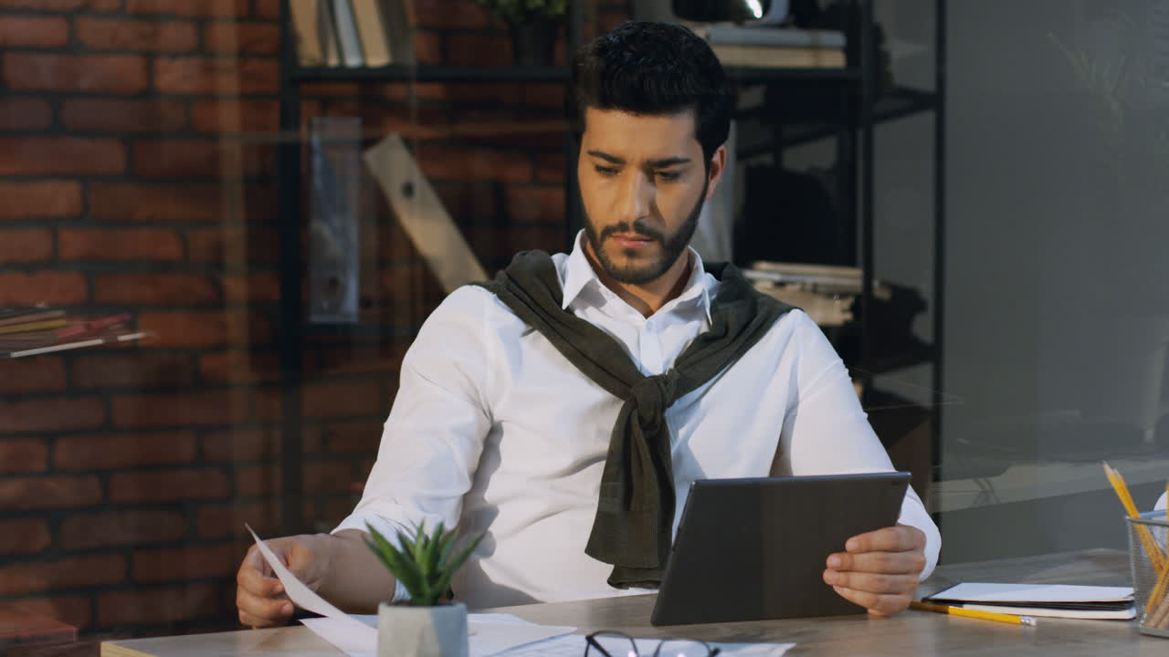 Young Arabian Businessman Sitting In His Office At The Desk And Working While Comparing Data Of The Document And On The Tablet