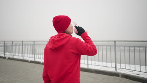 lady in red hoodie and beanie sips water from pink bottle outdoors in serene winter setting with snow, iron railing, and bridge in distant misty background