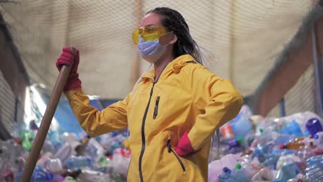 Portrait-Of-A-Girl-With-Dreadlocks,-In-Yellow-Jacket-And-Mask,-Holding-Large-Duck-Shovel,-Standing-At-A-Plastic-Recycling-Factory-1