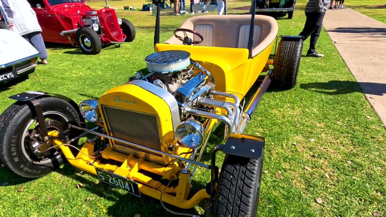 Bright yellow vintage hot rod with exposed engine viewed in sunlight, camera panning slowly across grassy outdoor car show in Tregony, Queensland