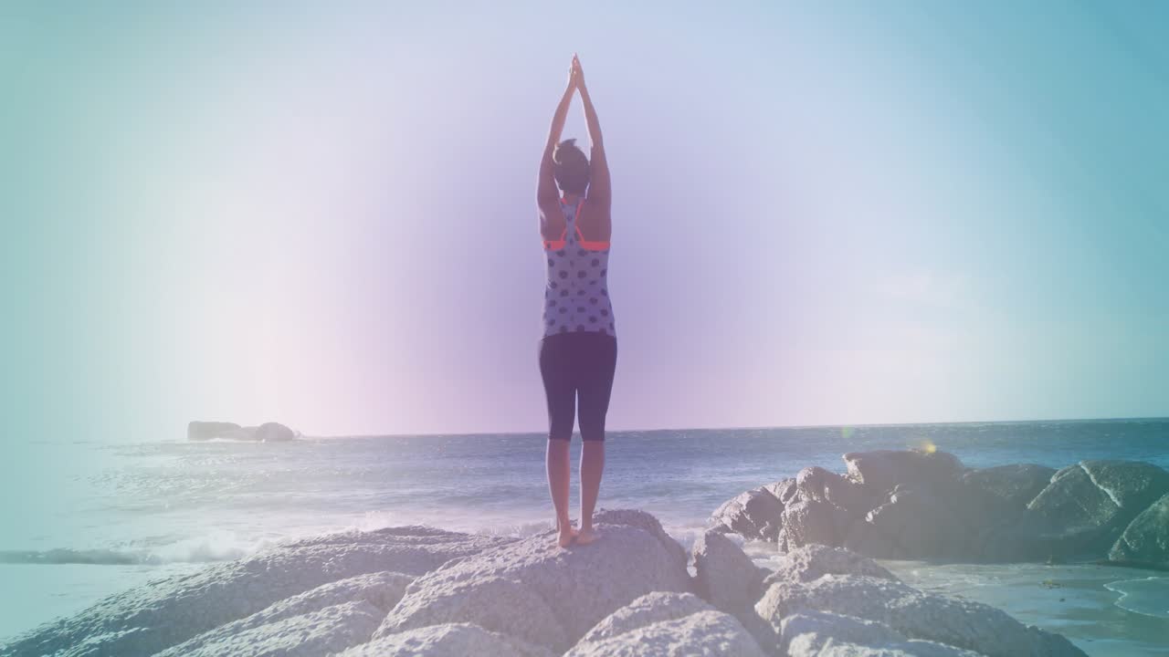 Donna che sale su una roccia centrale ed esegue un esercizio di stretching per l'equilibrio contro l'orizzonte dell'oceano.