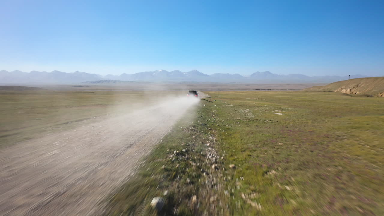 Ripresa con drone di un veicolo su una strada sterrata che guida vicino al lago Kel-Suu in Kirghizistan, partendo dalla nuvola di polvere dietro il SUV e rivelando un uomo che si sporge dal finestrino.