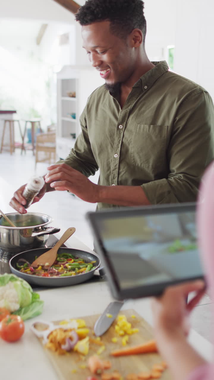 video vertical de una feliz pareja afroamericana con tableta cocinando en la cocina