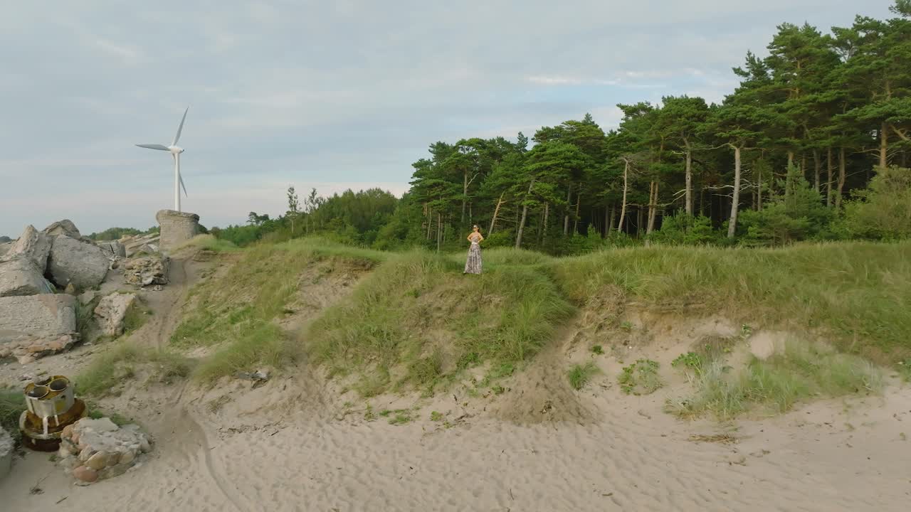vista aérea estableciendo de una hermosa joven romántica chica caucásica en un vestido largo en la playa de arena blanca, generador de viento en el fondo, soleada noche de verano, puesta de sol, amplia toma de drone moviéndose hacia atrás