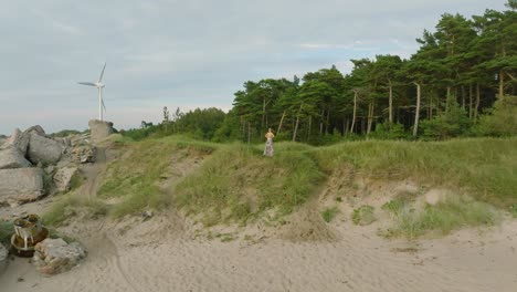 aerial establishing view of beautiful young romantic caucasian girl in a long dress on the white sand beach, wind generator in background, sunny summer evening, sunset, wide drone shot moving back