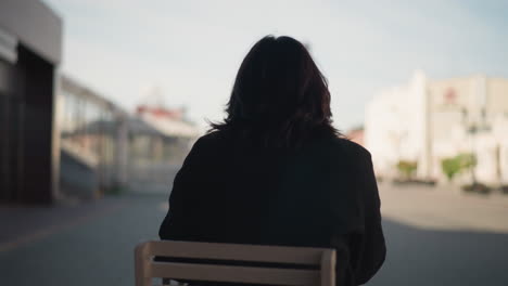 back view of lady in black chic coat seated on wooden chair outdoors, with blurred background featuring greenery, modern buildings, and soft natural light under an open sky in urban setting