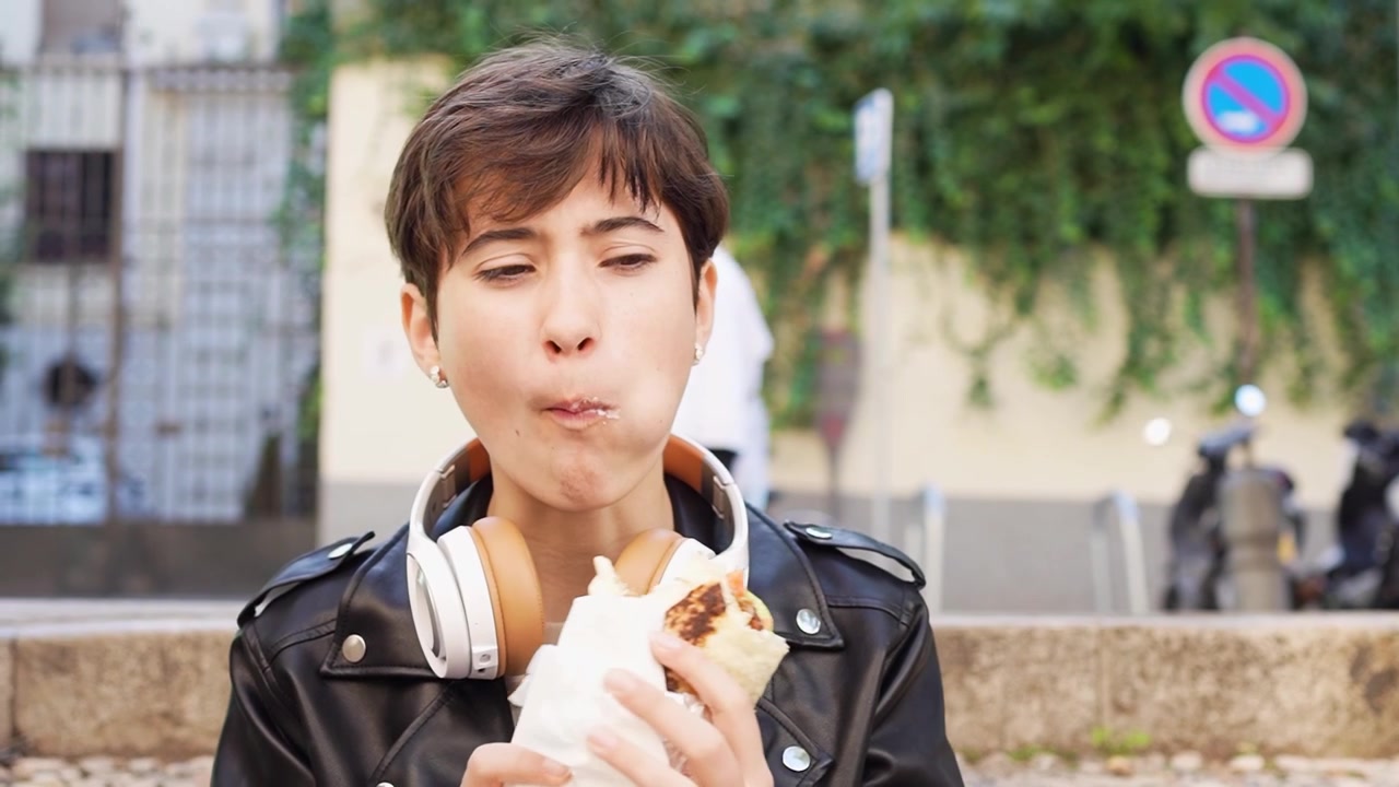 A young woman is happily enjoying her food in an urban setting while wearing headphones