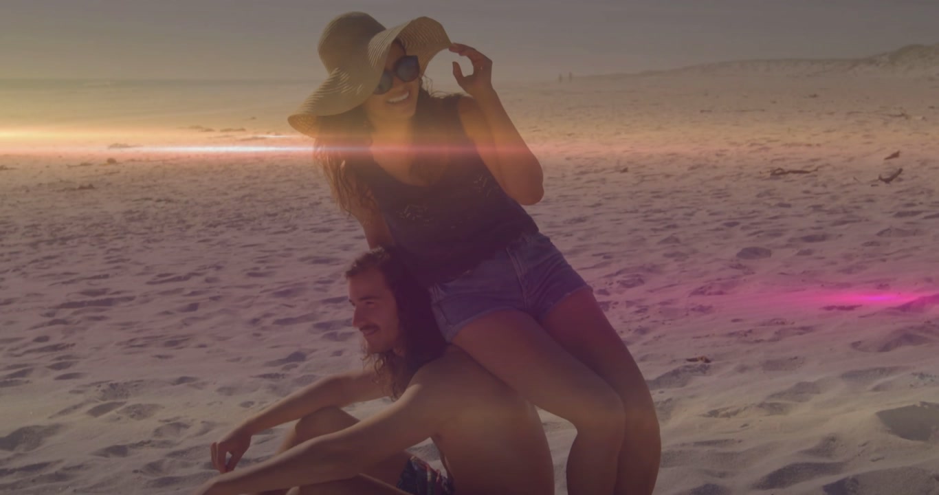Woman adjusting hat, shifting behind seated man on sunset beach, lens flare aiding travel marketing