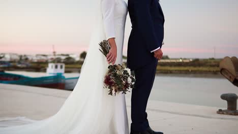 Close-up-of-married-bride-with-bouquet-of-flowers-and-groom-posing-next-to-a-lake-at-sunset